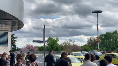 La-Gente-Que-Espera-En-El-Aeropuerto-De-Taxis-Recoge-El-Carril-Mientras-Un-Enorme-Avión-Militar-Se-Acerca-Al-Suelo-En-Un-Fondo-Nublado