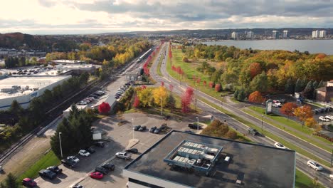 Las-Imágenes-De-Un-Dron-Capturan-Una-Vista-Aérea-De-Un-Paisaje-Urbano-Rebosante-De-Tráfico-Y-Actividad,-Con-Una-Carretera-Muy-Transitada,-Edificios-Comerciales-Y-Follaje-Otoñal-Bañado-Por-La-Suave-Luz-Del-Sol.