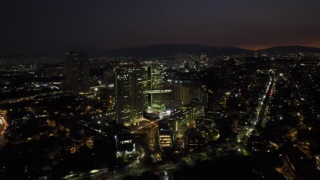 Vista-Aérea-Acercándose-Al-Complejo-Arcos-Bosques-Iluminado-Por-La-Noche,-En-La-Ciudad-De-México