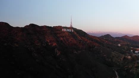 Toma-Aérea-De-Retroceso-Inverso-Del-Cartel-De-Hollywood-Al-Atardecer-Con-Montañas-Cubiertas-De-Nieve-En-El-Fondo