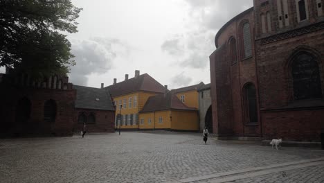 Slow-motion-dolly-shot-approaching-The-Absalon-Arch-between-Roskilde-Cathedral-and-the-Museum-of-Contemporary-Art-with-people-walking-by-in-Roskilde,-Denmark