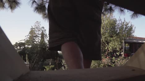 Young-hippie-man-walks-barefoot-down-the-wooden-stairs-which-take-him-to-a-green-garden,-slow-motion