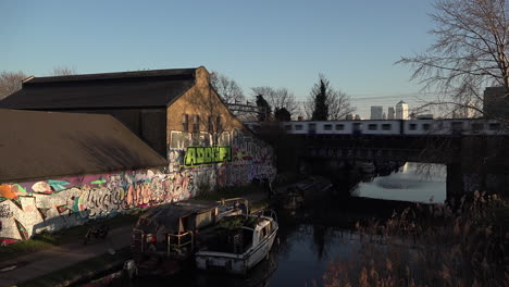 Reino-Unido-Febrero-De-2019:-Un-Tren-Cruza-Un-Puente-Sobre-El-Canal-Regents-Y-Una-Pared-Con-Grafitis-Junto-A-Un-Camino-De-Sirga-Con-El-Distrito-Financiero-De-Canary-Wharf-Al-Fondo