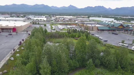 Imágenes-Aéreas-De-Un-Estanque-En-El-Estacionamiento-De-Cabela-En-Anchorage,-Alaska,-Que-Muestran-Muchos-Vehículos-Recreativos-Con-Autos-En-Movimiento,-Gente-Caminando,-Pájaros-Volando-Y-Montañas-En-El-Fondo.