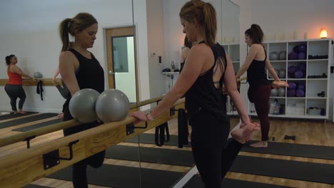 Damas-Trabajando-Frente-Al-Espejo-En-Un-Gimnasio-De-Yoga,-Usando-Traje-De-Ejercicio,-Un-Estante-De-Pelota-De-Ejercicio-En-La-Esquina