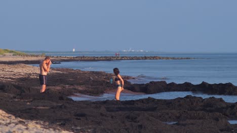 Young-couple-taking-pictures-at-beach-with-washed-up-dead-seaweed-in-the-evening-before-sunset,-medium-shot-from-a-distance