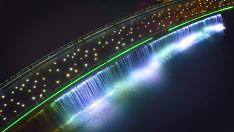 Luftaufnahme-Von-Links-Nach-Rechts,-Von-Oben-Nach-Unten,-Blick-Auf-Die-Starlight-Bridge-Oder-Anh-Sao-Bridge-Bei-Nacht,-Eine-Fußgängerbrücke-Mit-Farbigen-Lichtern-Und-Wasserfall-Im-Bezirk-7-Von-Ho-Chi-Minh-Stadt-Oder-Saigon,-Vietnam