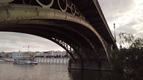 Tiro-Inclinado-Hacia-Abajo-Desde-El-Icónico-Puente-De-Triana-Hasta-El-Paseo-En-Barco-Por-El-Río-En-Sevilla,-España