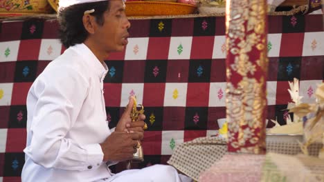 Hindu-priest-dressed-in-white-holding-religious-item-prays-at-a-colorfully-decorated-altar-of-a-temple-in-Bali,-Indonesia