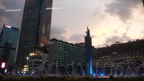 Slow-motion-shot-circling-around-the-circular-park-with-water-splashing-up-in-a-perfect-circle-around-the-monument-that-is-lit-up-by-blue-light-in-central-Jakarta,-Indonesia