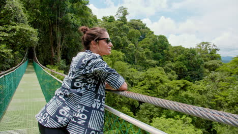 Moviéndose-En-Una-Foto-De-Una-Hermosa-Morena-Con-Gafas-De-Sol-Disfrutando-Del-Panorama-En-Un-Puente-Colgante-En-Medio-Del-Bosque-En-Arenal,-Costa-Rica