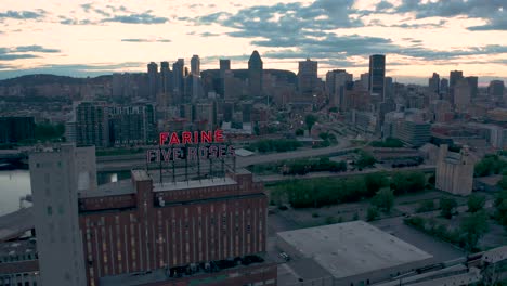 Drone-Volando-Lejos-Del-Famoso-Letrero-Rojo-Parpadeante-Del-Edificio-Farine-Cinco-Rosas,-Con-El-Centro-De-Montreal-En-El-Fondo