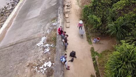 Mujer-Vietnamita-Con-La-Cara-Cubierta-Moviendo-Una-Carretilla-Con-Grava-Pasando-A-Algunos-Turistas-Masculinos,-Vista-Superior-Panorámica-Hacia-Abajo