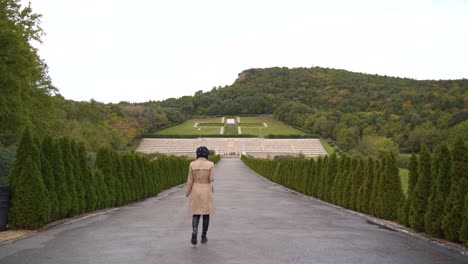 View-of-female-tourist-entering-shot-at-Polish-cemetery-at-Monte-Cassino's-asphalt-walkway-lined-with-green-trees,-Italy,-static