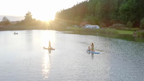 A-wide-drone-shot-of-a-family-paddle-boarding-on-a-lake-during-sunset-at-Mountain-Petzen-Pirkdorfer-See,-Austria