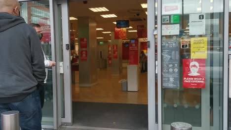 People-standing-in-front-of-the-shop-with-face-masks-with-warning-signs-about-distance-and-face-masks-on-doors