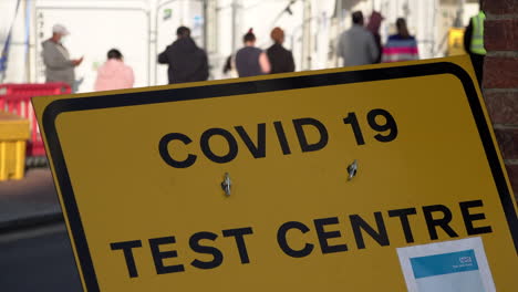 People-in-protective-face-masks-queue-up-outside-an-appointment-only-Covid-19-testing-centre-behind-a-yellow-Covid-19-Testing-Centre-street-sign