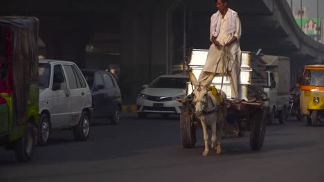 Un-Coche-De-Burros-Va-Por-La-Carretera,-El-Tráfico-Se-Cierra-En-La-Carretera-Bajo-El-Sobrevuelo,-Pasando-Motos-Y-Coches,-Autobuses,-Rickshaw,-Vallas-Publicitarias-Y-Edificios-En-El-Fondo