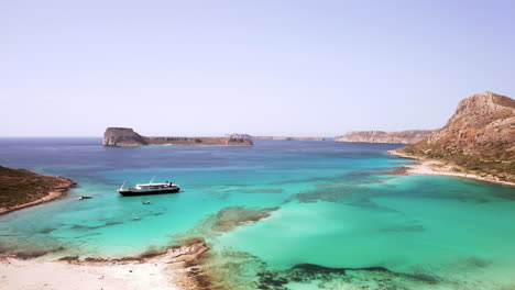 Aerial-Flyby-of-Day-Cruise-Boat-Departing-Balos-Beach-in-Crete,-Greece-on-Beautiful-Sunny-Day-with-Turquoise-Water---White-Sand