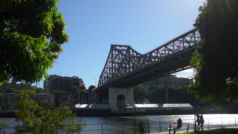 Los-Caminantes-Matutinos-Pasean-Por-La-Orilla-Del-Río-Brisbane,-Bañado-Por-El-Sol,-En-Kangaroo-Point,-Con-El-Icónico-Puente-Story-Elevándose-En-Lo-Alto