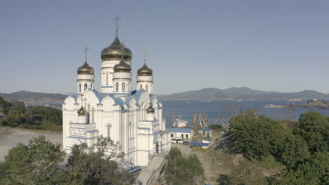 Toma-Aérea-De-Descenso-Lento-De-La-Iglesia-Ortodoxa-Junto-Al-Mar-Con-Techo-Azul-Y-Cúpulas-Doradas-Con-Puerto-Y-Bahía-En-El-Fondo-En-Un-Día-Brillante-Y-Soleado