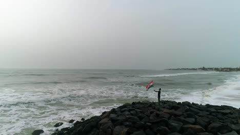 Grupo-De-Hombres-Jóvenes-Patriotas-Sostienen-La-Bandera-Nacional-India-De-Pie-En-Una-Playa-Rocosa-Con-Olas-Rompiendo