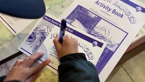 Niño-Escribiendo-Su-Nombre-En-Un-Certificado-De-Guardabosques-Junior-En-El-Parque-Nacional-Del-Monte-Rainier-Y-También-Recibiendo-Una-Insignia-De-Oro