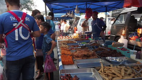 Puestos-De-Comida-Llenos-De-Comida-En-La-Mesa-Con-Gente-Reunida-Y-Comprando-Mientras-El-Vendedor-Está-A-La-Espera