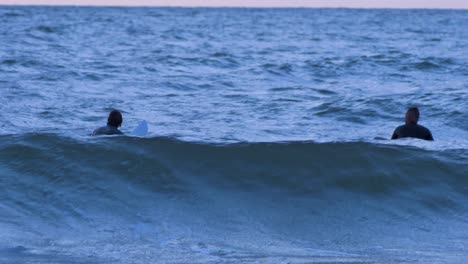 Surfers-on-surfboards-paddling-over-waves-near-the-Baltic-sea-Karosta-beach-at-Liepaja-during-a-beautiful-vibrant-sunset-at-golden-hour,-medium-shot-from-a-distance