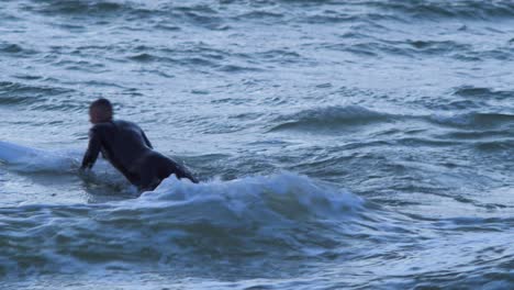 Surfer-on-surfboard-paddling-over-waves-near-the-Baltic-sea-Karosta-beach-at-Liepaja-during-a-beautiful-vibrant-sunset-at-golden-hour,-medium-shot-from-a-distance