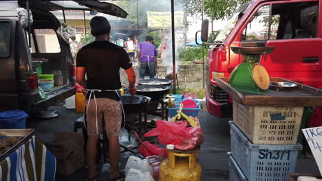 Zwei-Männer-Kochen-Essen-Auf-Drei-Großen-Woks,-Umgeben-Von-Einem-Roten-Lastwagen,-Einem-Benzintank,-Einer-Waage-Und-Plastikbehältern