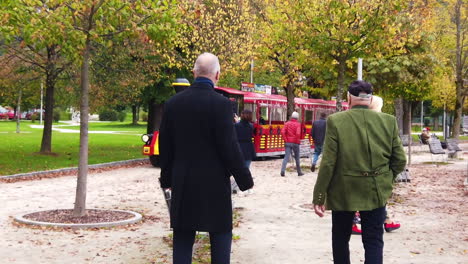 Two-men-looking-like-a-gangster-with-a-blond-lady,-a-fun-train-and-chairs-in-the-park