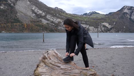 Mujer-Con-Auriculares-Toma-Un-Descanso-De-Trotar-Para-Atar-El-Zapato-En-El-Lago-De-Montaña