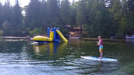 Carro-Aéreo-Hacia-Atrás,-Un-Niño-Observa-Desde-Una-Tabla-Flotante-En-Un-Lago-Mientras-Otros-Niños-Saltan-De-Un-Juguete-Inflable-&quot;blob&quot;-Y-Se-Lanzan-Unos-A-Otros-Al-Aire