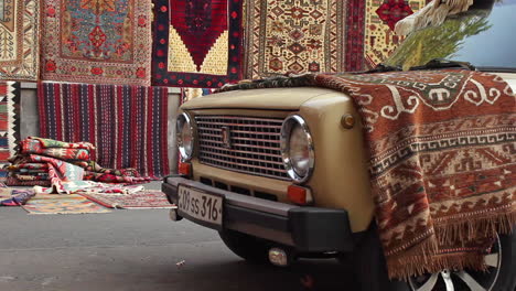 Coche-Aparcado-Con-Una-Alfombra-De-Colores-Sobre-El-Capó-Contra-El-Fondo-De-Una-Alfombra-Colgante-Hecha-A-Mano