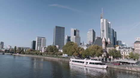 Statische-Aufnahme-Der-Frankfurter-Skyline-Mit-Wolkenkratzern-Und-Fähre-Im-Vordergrund,-Blick-Vom-Hauptfluss-An-Einem-Sonnigen-Tag-Mit-Klarem-Himmel,-Hessen,-Deutschland