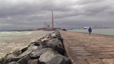 Algunas-Olas-A-La-Izquierda-Y-Dublin-Esb-Poolbeg-En-El-Centro-Con-Una-Tienda-A-La-Derecha,-Algunas-Personas-Caminando-A-Lo-Largo-De-La-Costa-O-Pared-Sur