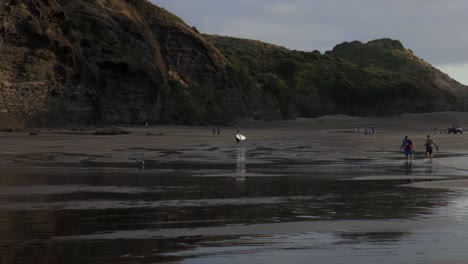 Surfer-Läuft-Mit-Surfbrett-Ins-Meer-Bei-Sonnenuntergang-Am-Strand-Von-Piha,-Piha,-Nordinsel,-Neuseeland