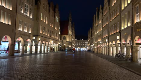 Wide-shot-of-illuminated-old-town-with-old-arch-buildings,-shops-amd-riding-cyclist-on-the-road-during-night