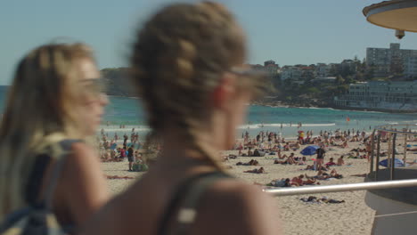 Una-Concurrida-Playa-De-Bondi-Con-Un-Par-De-Mujeres-Que-Cruzan-La-Cámara-Y-Revelan-La-Casa-De-Los-Salvavidas-En-Sydney,-Australia