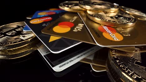 Cerrar-Girando-Bitcoins-Btc-Y-Tarjetas-De-Débito-De-Crédito-Mastercard-En-Superficie-De-Vidrio-Reflectante-Negro,-Pagos-De-Criptomoneda-4k