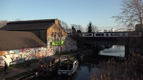 Reino-Unido-Febrero-De-2019--Tres-Trenes-Se-Cruzan-Entre-Sí-En-El-Puente-Sobre-El-Canal-Regents-Y-Una-Pared-Pintada-Junto-A-Un-Camino-De-Sirga-Con-El-Distrito-Financiero-De-Canary-Wharf-En-El-Fondo