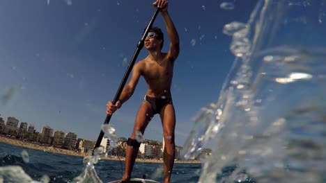 Joven-Remando-De-Pie-Con-La-Playa-De-La-Ciudad-En-El-Fondo