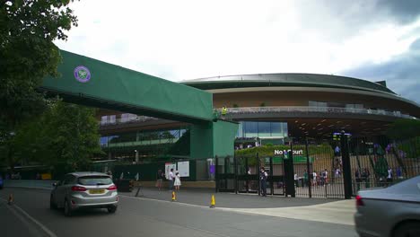 Vista-De-La-Entrada-De-Wimbledon-Con-La-Cancha-Número-1-Al-Frente,-Automóviles-Y-Personas-Que-Pasan,-El-Puente-Peatonal-Se-Usa-Para-Conectar-La-Cola-A-Las-Distintas-Canchas