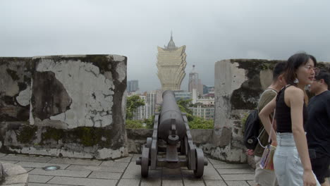 Touristen,-Die-An-Der-Alten-Metallkanone-Auf-Der-Fortaleza-Do-Monte-Vorbeikommen-Und-In-Richtung-Hotel-Grand-Lisboa-In-Macau,-Macau-Sar,-China-Zeigen