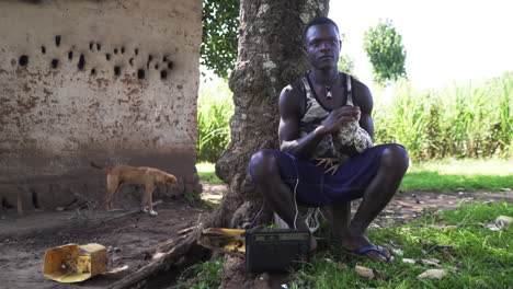 Joven-Sentado-En-Una-Silla-Pequeña-Mientras-Acaricia-Un-Pollo-Y-Escucha-La-Radio-Con-Un-Perro-Que-Pasa-Al-Fondo,-Uganda-áfrica