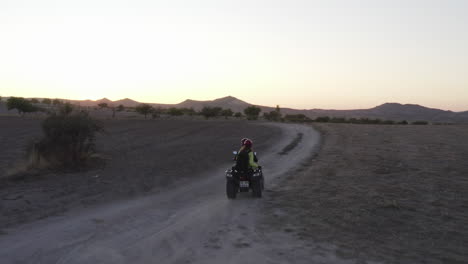 Drone-following-couple-on-ATV-quad-going-down-a-dusty-farm-road-in-Love-Valley-in-Goreme,-Turkey