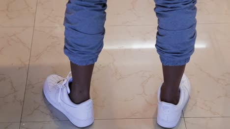 Primer-Plano-De-Las-Piernas-De-Un-Modelo-Masculino-Negro-Con-El-Dobladillo-Enrollado-De-Sus-Pantalones-Azules-Usando-Zapatillas-Blancas-Haciendo-Un-Giro-De-360-Grados-En-Un-Piso-Beige---Disparo-De-Trípode-De-Escena-Interior