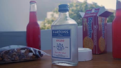 Handheld-Moving-Wide-Shot-of-a-Bottle-of-Ouzo-With-Nuts,-Digestive-and-Some-Bottles-of-Margarita-in-The-Background-With-Moving-Tree-By-The-Wind