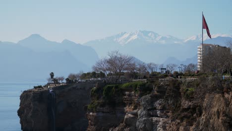 Plano-Completo,-Turista-Del-Acantilado-De-Antalya-Caminando,-Vista-Panorámica-De-Las-Montañas-De-Toros-En-El-Fondo,-Bandera-De-Turquía-En-El-Costado